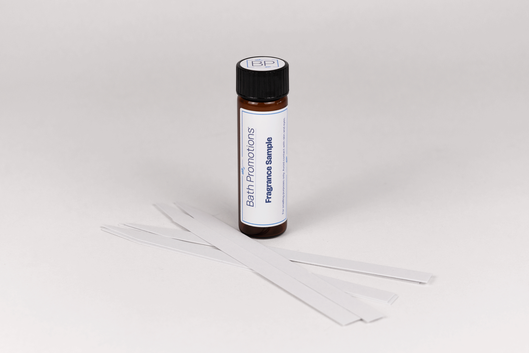 An amber glass vial labeled with a Fragrance Sample Bath Promotions branded label standing upright with paper scent blotters in front of it.