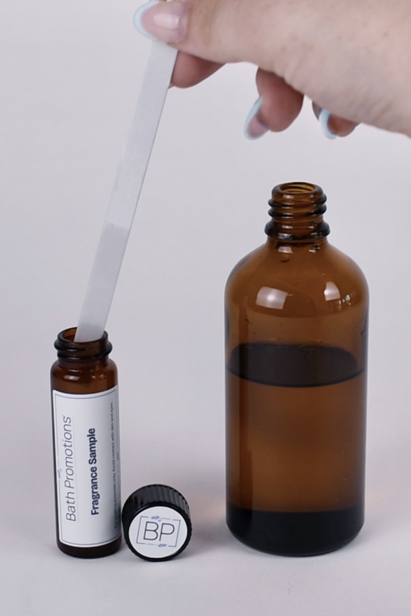 A scent blotter infused with fragrance being placed inside a protective amber glass vial labeled with Bath Promotions branding next to a large amber glass bottle of fragrance oil.