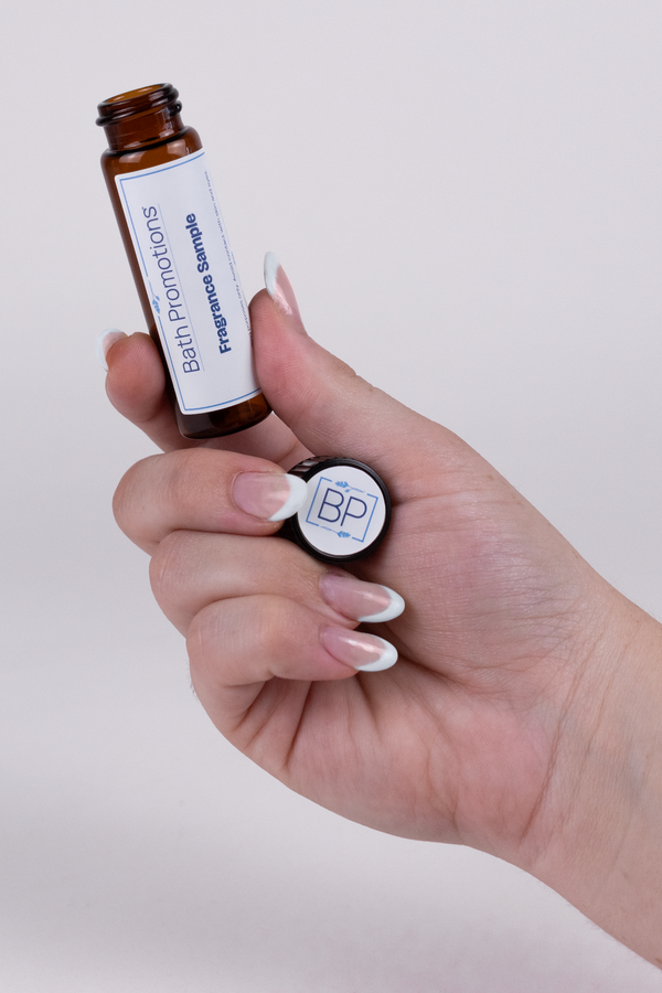 An amber glass vial labeled with a Bath Promotions branded label being held by a hand of light complexion and a white French tipped manicure.