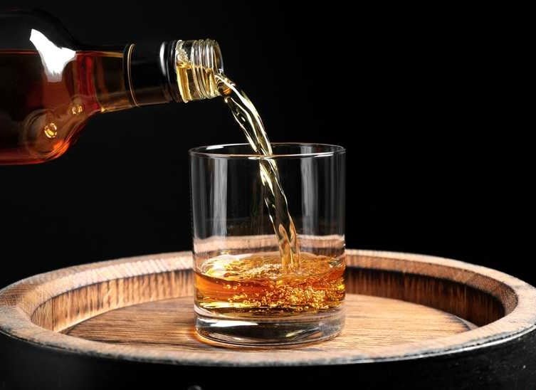 Amber whiskey being poured from bottle into rocks glass on wooden barrel top against black background.
