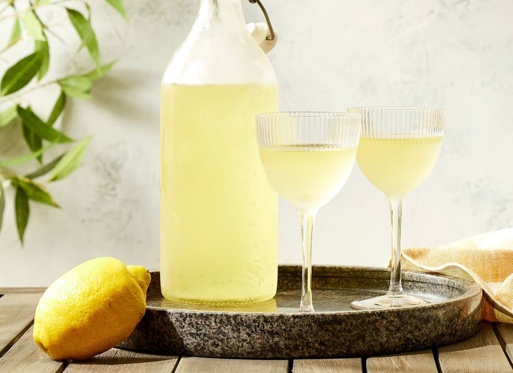 Yellow limoncello in glass bottle and two wine glasses on stone tray with fresh lemon and green leaves.