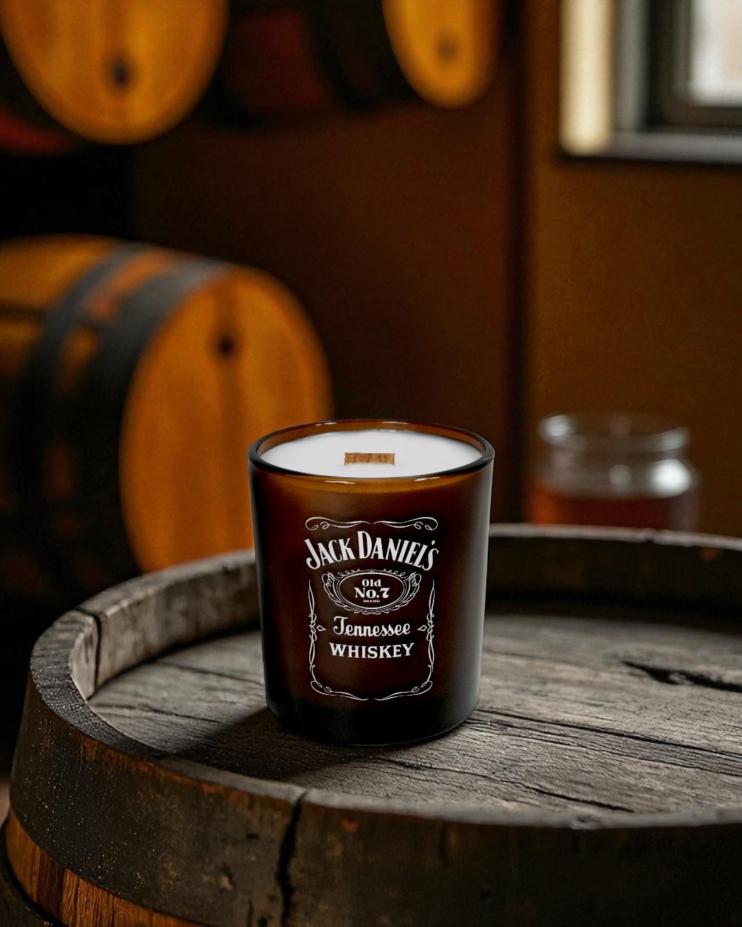 A amber 14 oz. glass candle with wooden wick featuring Whiskey logo sits on a weathered wooden whiskey barrel, with additional barrels visible in the blurred distillery background