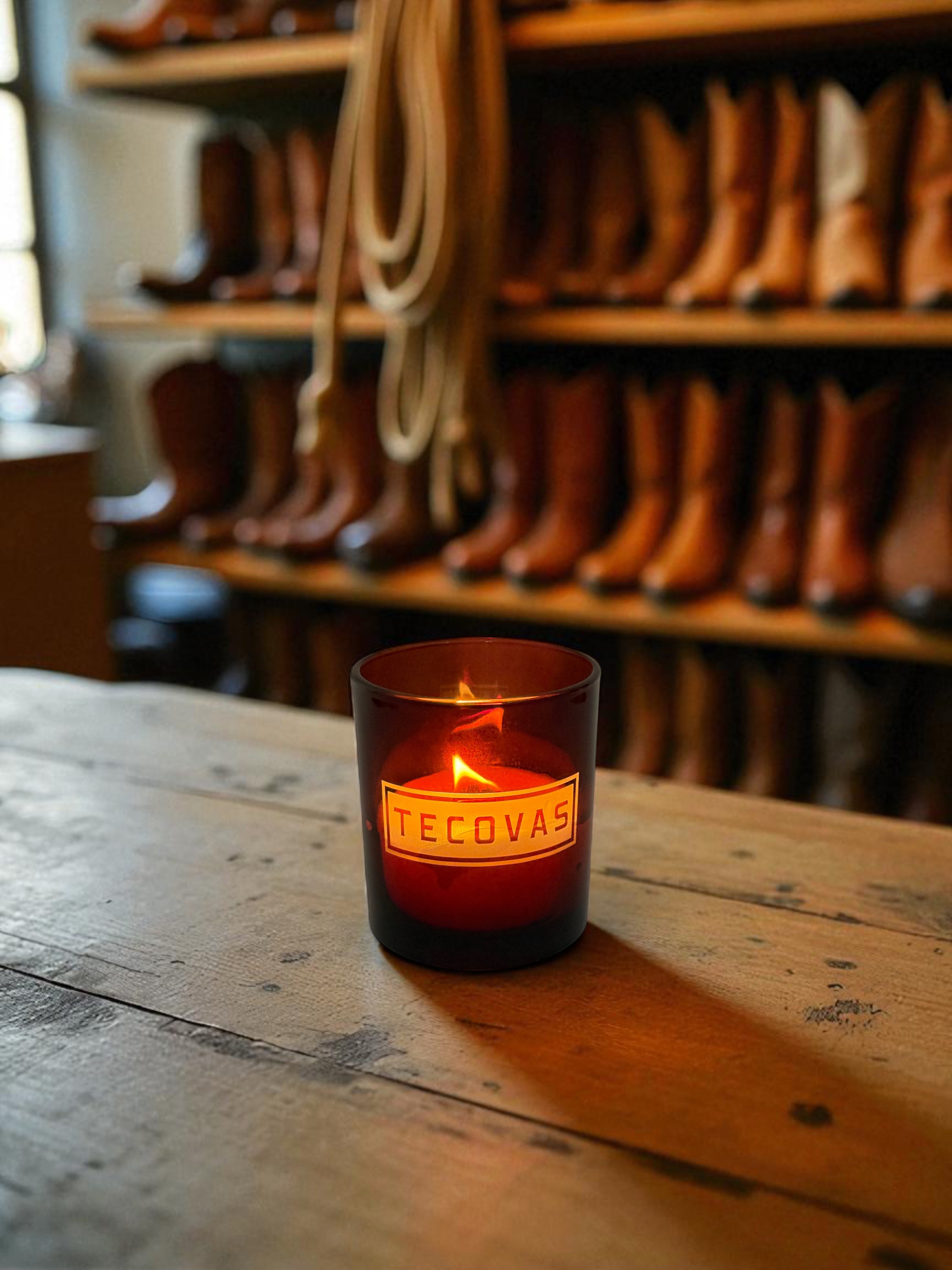 A lit amber 14 oz. glass candle with engraved text sits on a weathered wooden surface in a western boot store, with rows of leather cowboy boots displayed on wooden shelves in the background
