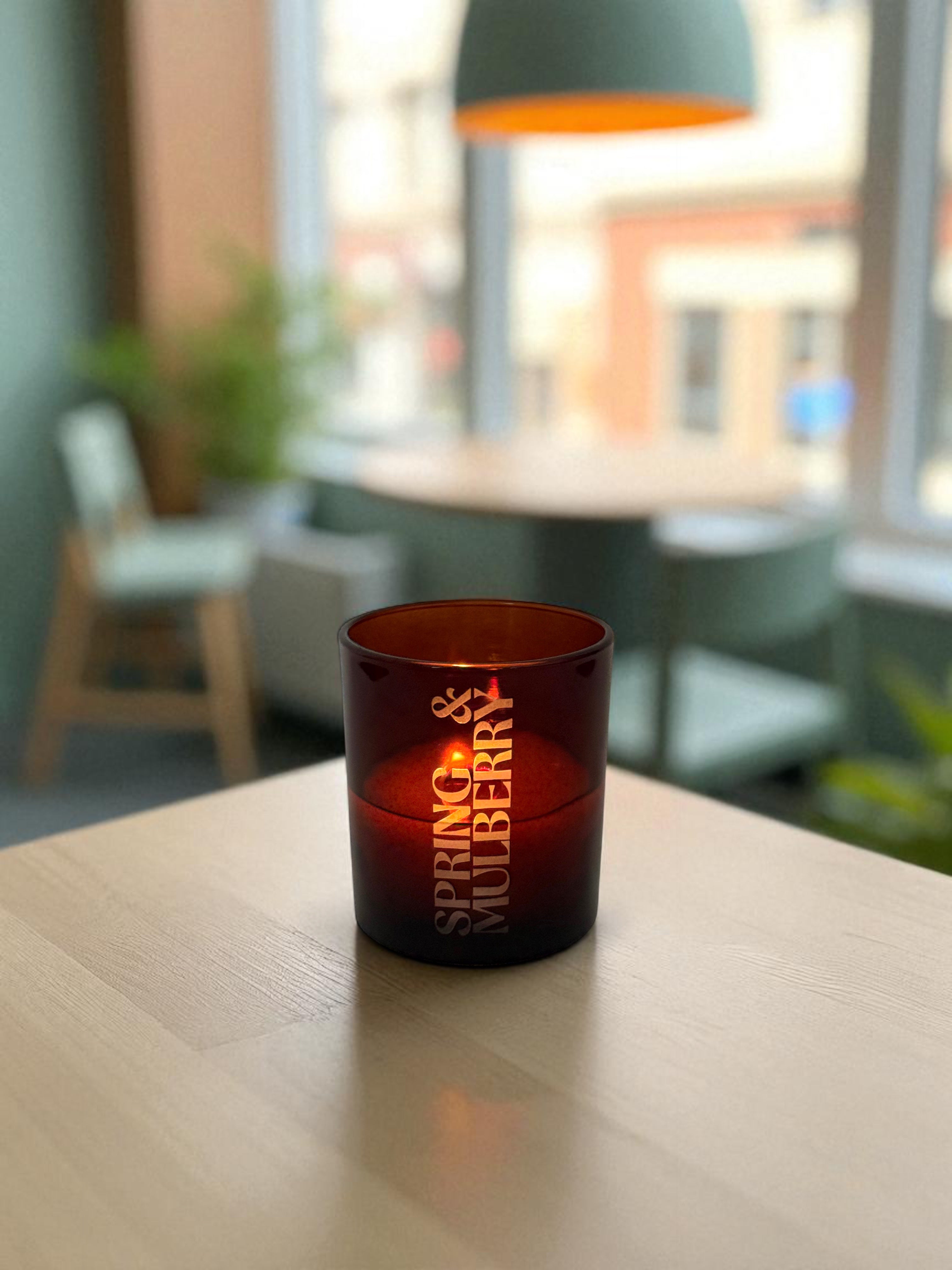 A lit 14 oz. amber glass candle with engraved text sits on a light wood table in a modern cafe, with blurred seating area and large windows showing street view in the background