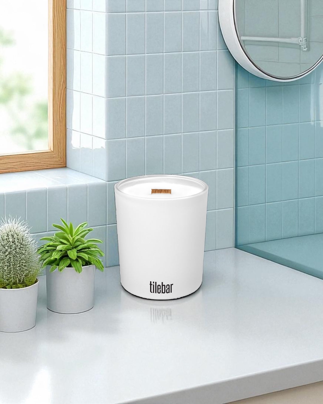 A white 14 oz. candle with wooden wick and black text sits on a white bathroom countertop next to small potted plants, with light blue subway tiles, a round mirror, and wooden window frame in the background