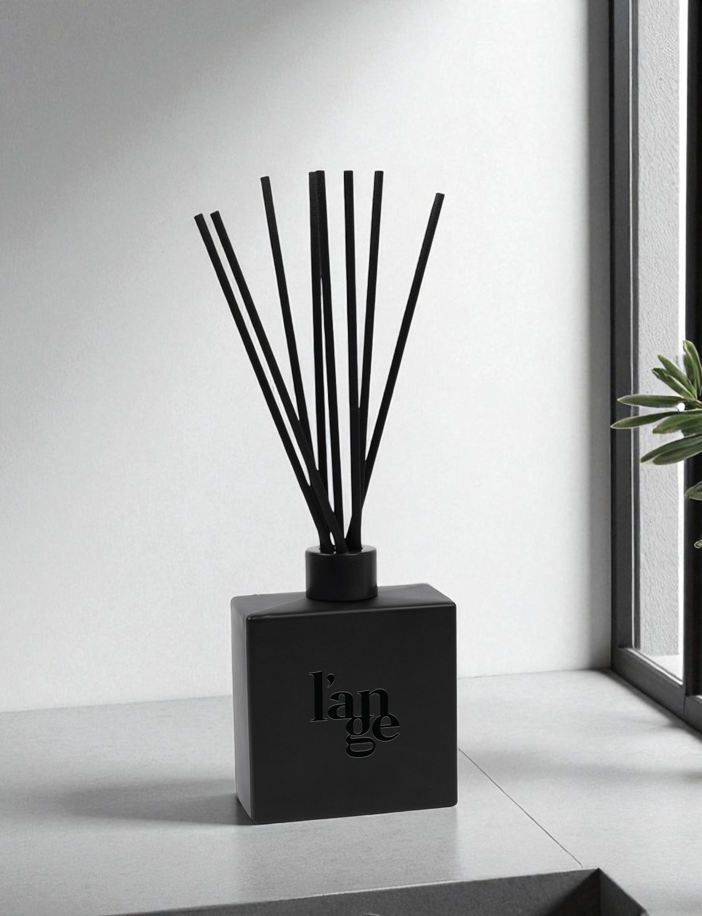 A matte black greed diffuser with glossy text and black reed sticks sits on a white windowsill in a minimalist modern interior, with natural light streaming through the window and a small green plant visible in the corner