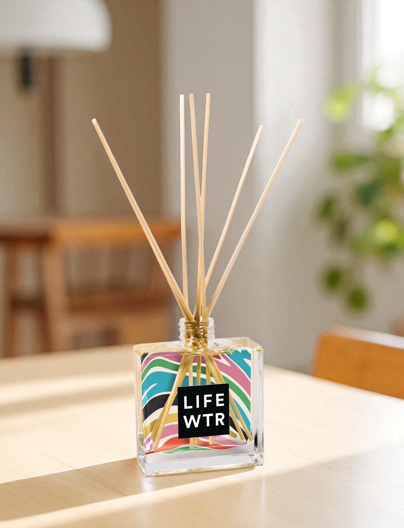 A clear glass reed diffuser with colorful pattern and text with wooden reed sticks sits on a wooden counter next to a plant at the distance