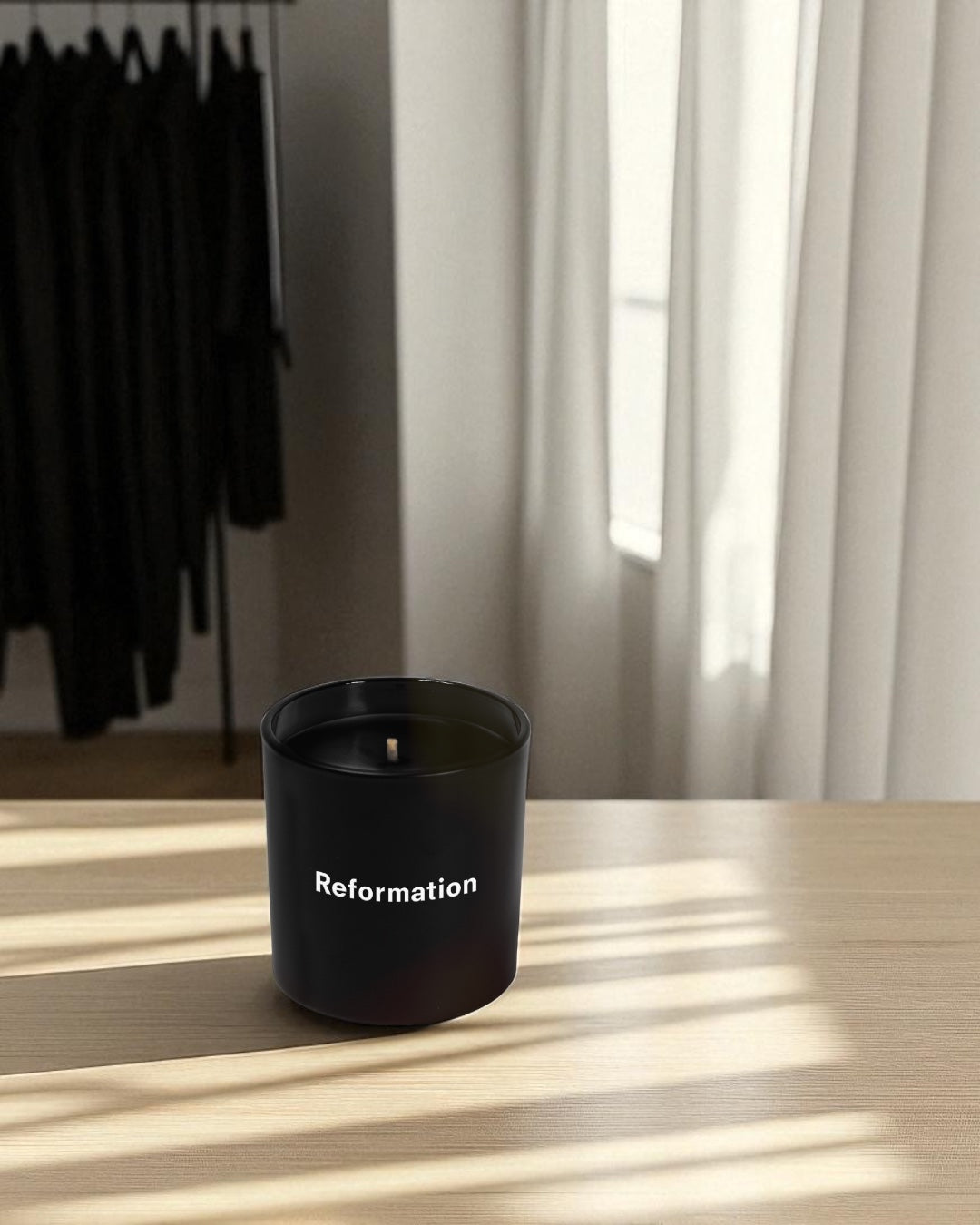 A black 9 oz. candle with white imprint sits on a light wooden surface in natural sunlight. The candle has a cotton wick and is positioned in a minimalist room with white curtains and black clothing hanging on a rack in the background.