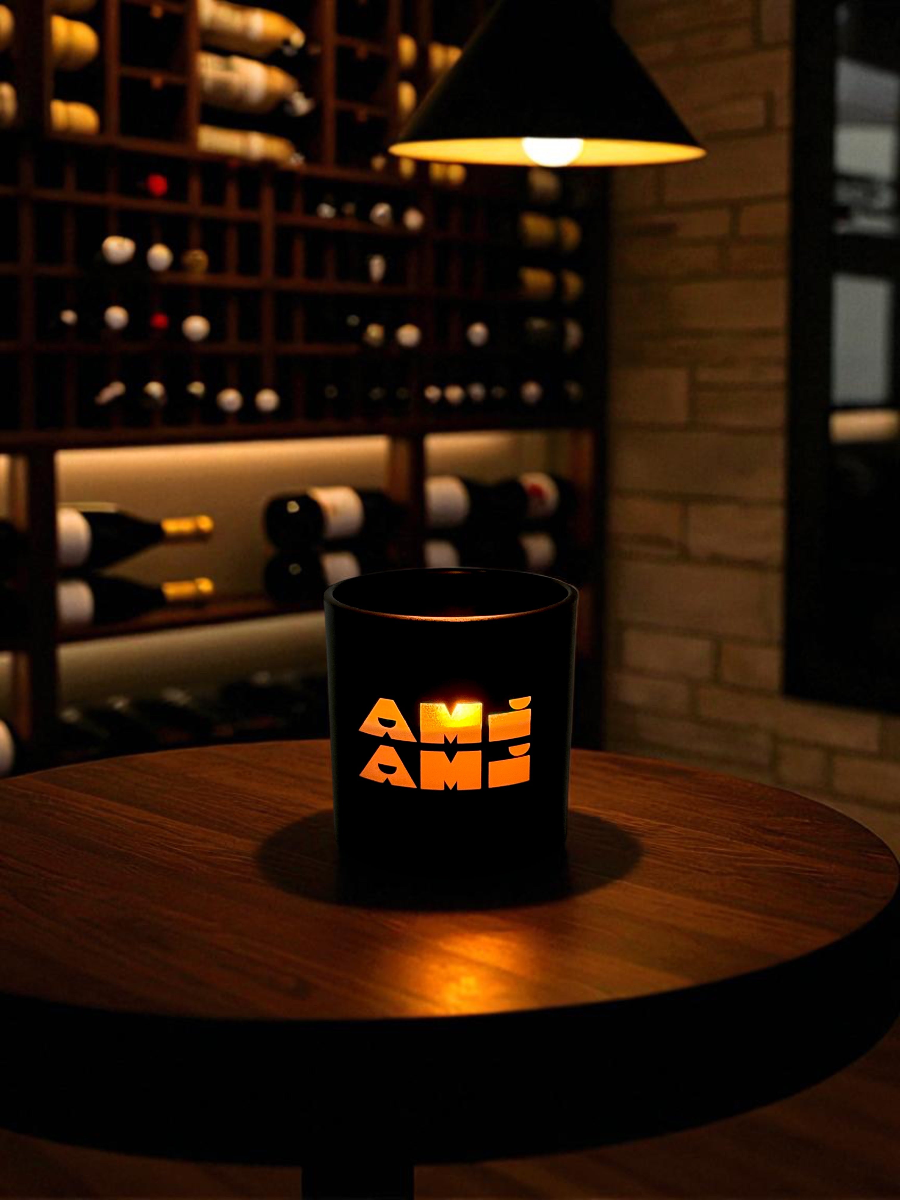 A black 9 oz. candle with engraved logo glows warmly on a round wooden table in an upscale wine cellar. Behind the candle, wine bottles are stored in wooden racks with ambient lighting, and a pendant light hangs overhead, creating an intimate dining atmosphere.