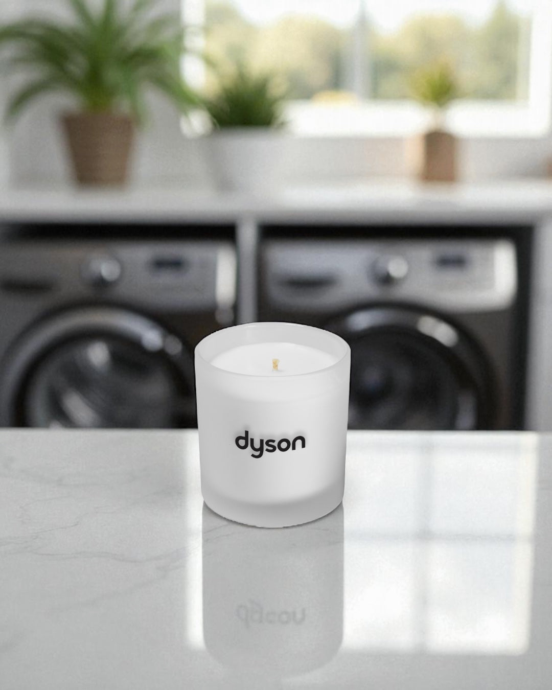 A frosted 9 oz. glass candle with black text sits on a reflective white surface in a modern laundry room. The candle has a cotton wick and is positioned in front of washer and dryer units, with potted plants visible on a shelf in the blurred background.