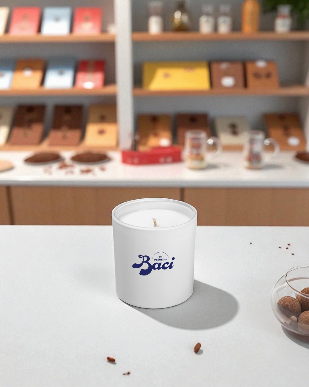 A white 9 oz. candle with blue logo text, sitting on a white surface with chocolate shelving display in the blurred background