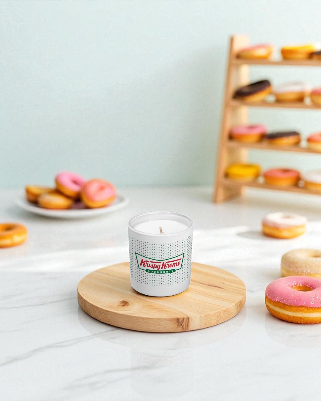 A white 9 oz. candle with green and red logo sits on a round wooden board surrounded by colorful glazed donuts on a marble countertop, with a wooden donut display rack in the background