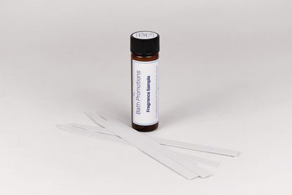 An amber glass vial labeled with a Fragrance Sample Bath Promotions branded label standing upright with paper scent blotters in front of it.