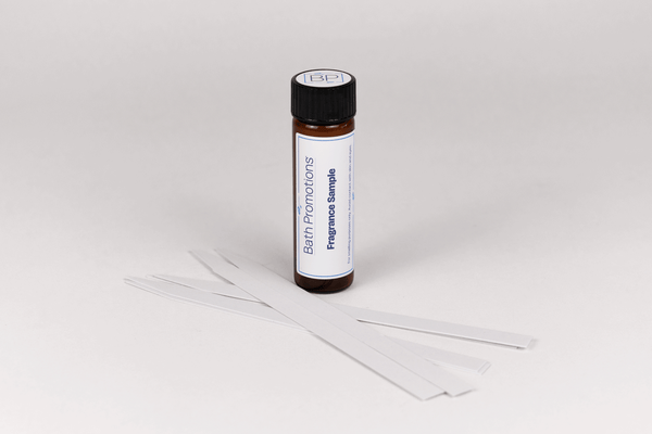 An amber glass vial labeled with a Fragrance Sample Bath Promotions branded label standing upright with paper scent blotters in front of it.