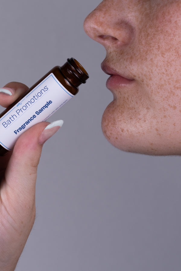 A person of light complexion and a white French tipped manicure holding up an amber glass vial labeled with a Bath Promotions branded label up to their face to enjoy the fragrance in the vial.