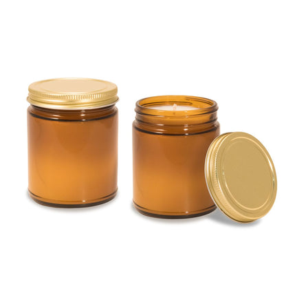 Two 8 oz amber glass apothecary jar candles with gold screw-on lids. One jar is closed with lid on top, the other is open showing the lit candle inside with the lid placed beside it