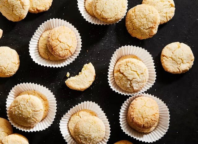Golden-brown amaretti cookies arranged on a dark surface, some placed in white paper cupcake liners and others scattered around them, with one cookie broken to show the crumbly interior texture.