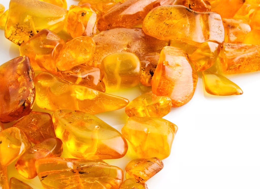 Collection of translucent amber-colored stones and crystals in various shapes and sizes, ranging from golden yellow to deep orange, scattered on a white background with natural lighting creating warm glows and reflections.