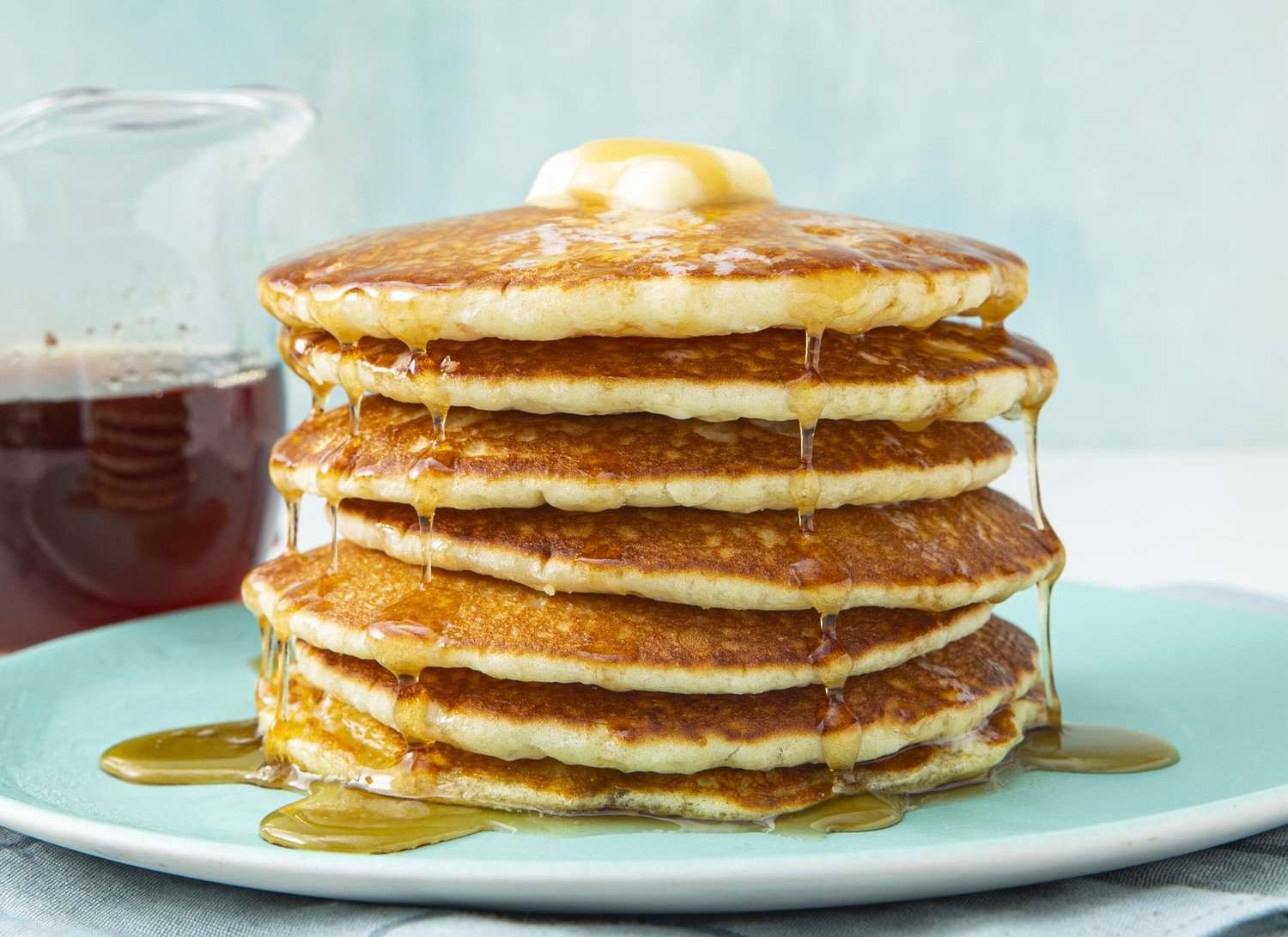 Stack of golden brown pancakes on white plate with butter pat on top and maple syrup dripping down sides, with glass pitcher of syrup in background.