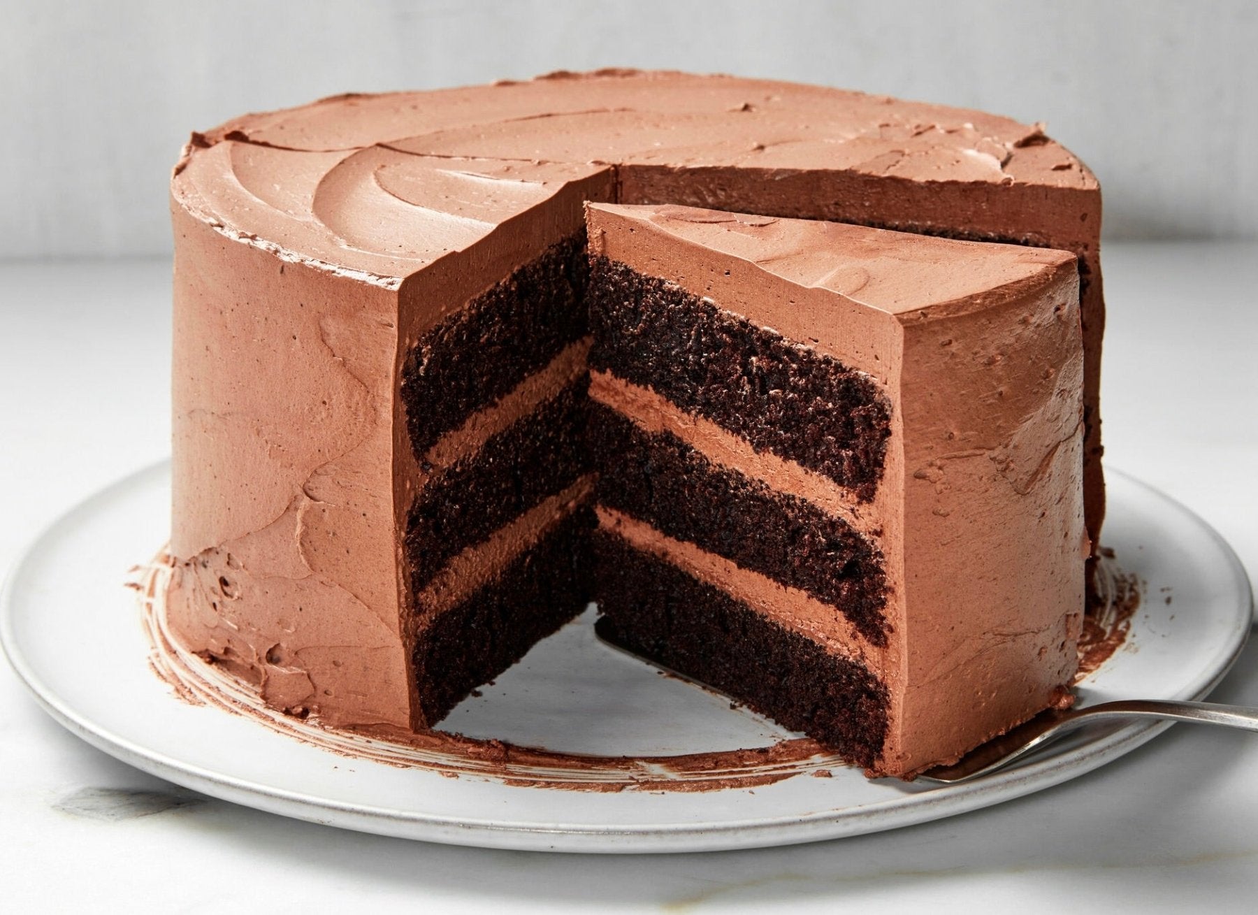 Three-layer chocolate cake with chocolate frosting on white plate, showing cross-section with dark chocolate cake layers and chocolate buttercream filling.