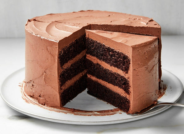 Three-layer chocolate cake with chocolate frosting on white plate, showing cross-section with dark chocolate cake layers and chocolate buttercream filling.