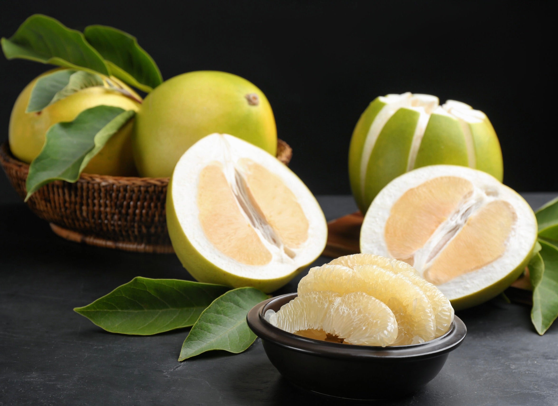 Fresh white grapefruit halves and segments with green leaves in wicker basket and black bowl on dark surface.