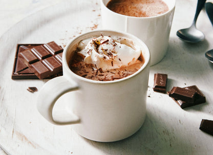 White mugs of hot chocolate with whipped cream and chocolate shavings, surrounded by chocolate pieces on wooden surface.