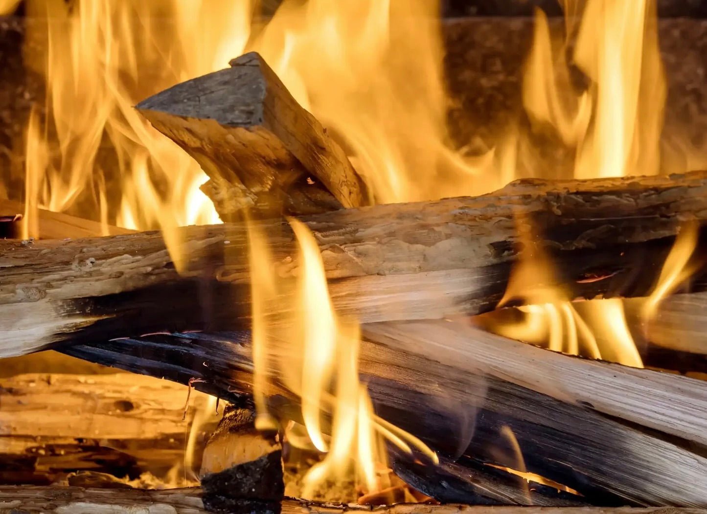 Bright orange flames rising from burning wooden logs in a fire with charred and weathered wood pieces.