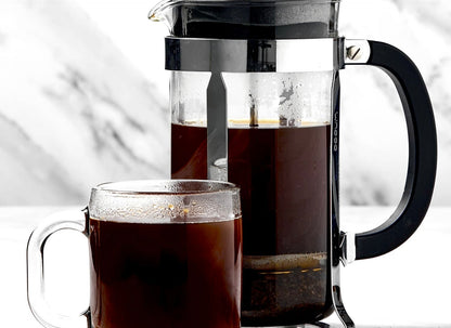 Glass French press coffee maker with dark coffee and clear glass mug filled with coffee on white surface.