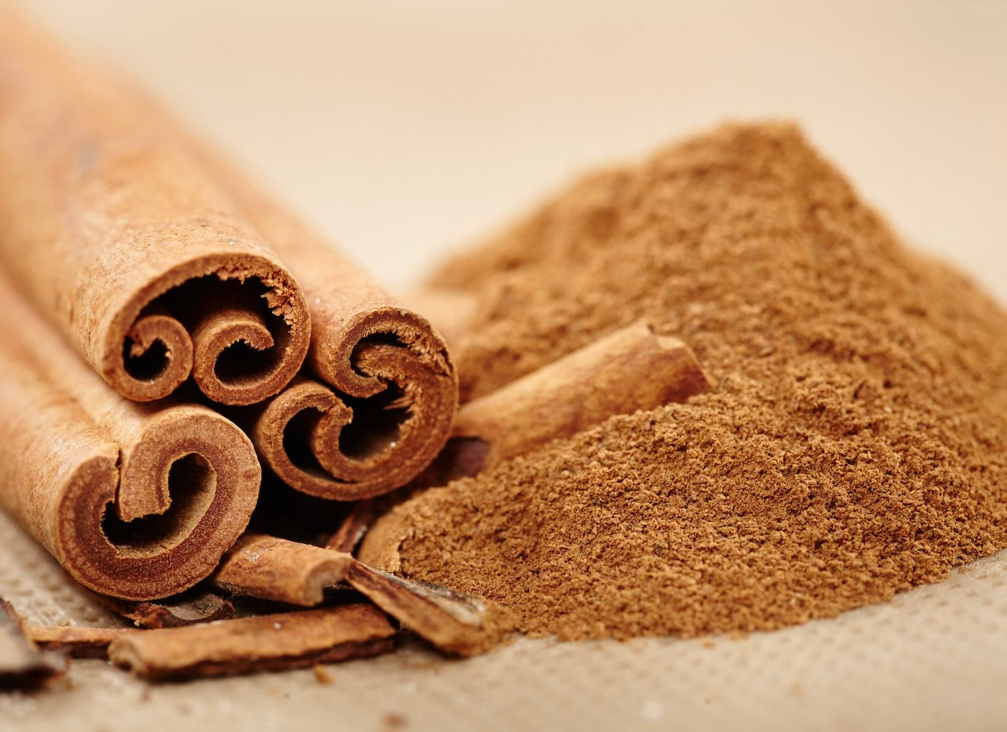 Cinnamon sticks with curled bark texture next to pile of fine brown ground cinnamon powder on light surface.