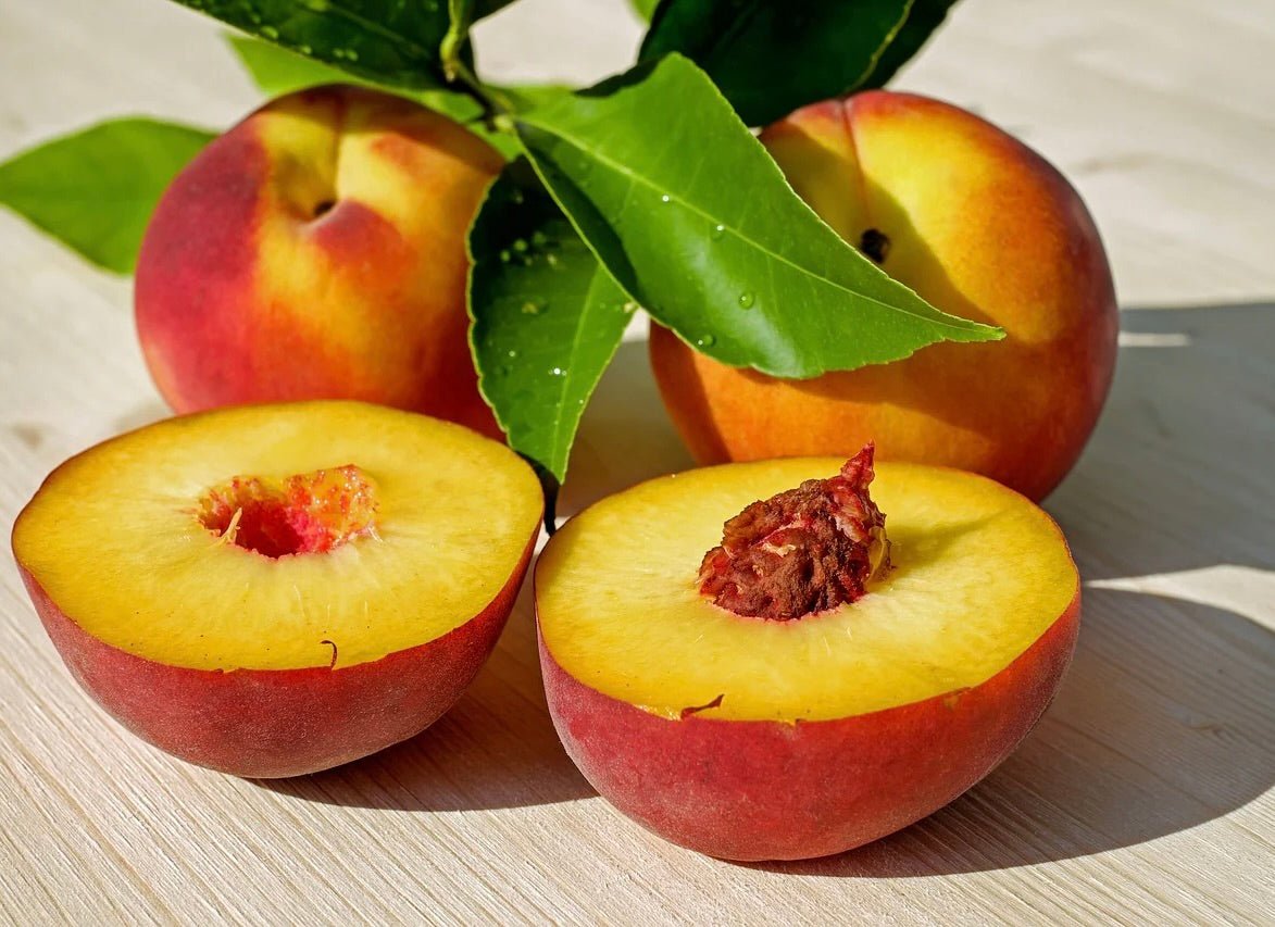 Fresh peaches with red and yellow skin, two halved showing golden flesh and brown pits, with green leaves on wooden surface.