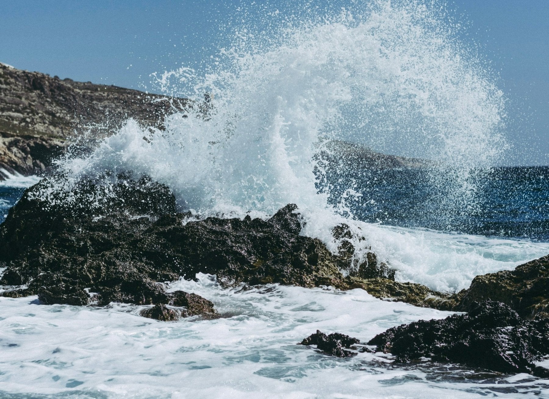 Ocean waves crashing against dark rocky coastline creating dramatic white water spray against blue sky.