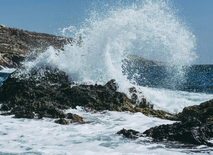 Ocean waves crashing against dark rocky coastline creating dramatic white water spray against blue sky.