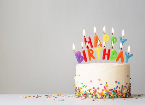 White birthday cake with colorful sprinkles and lit letter candles spelling HAPPY BIRTHDAY on gray background.
