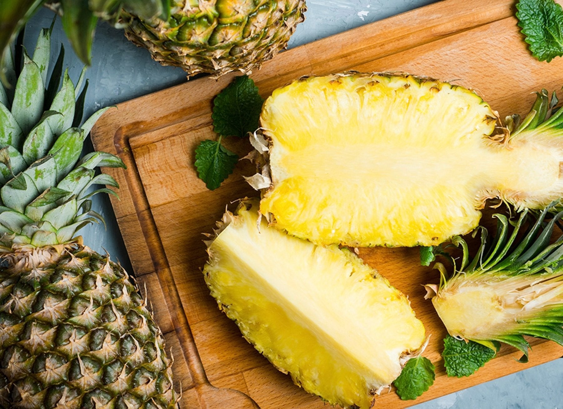 Fresh pineapples with one halved showing bright yellow flesh on wooden cutting board with green mint leaves.