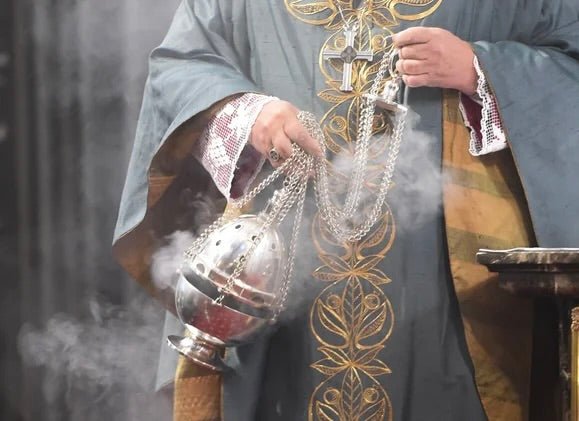 Person in ornate religious vestments holding silver incense burner with white smoke rising during ceremony.