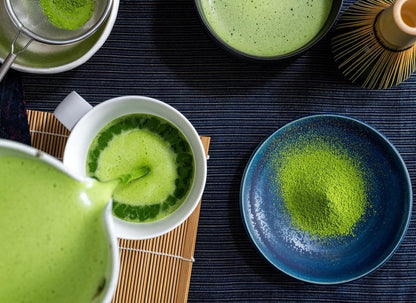 Japanese matcha tea ceremony setup with green tea in white cup, matcha powder in blue bowl, and bamboo whisk on dark mat.