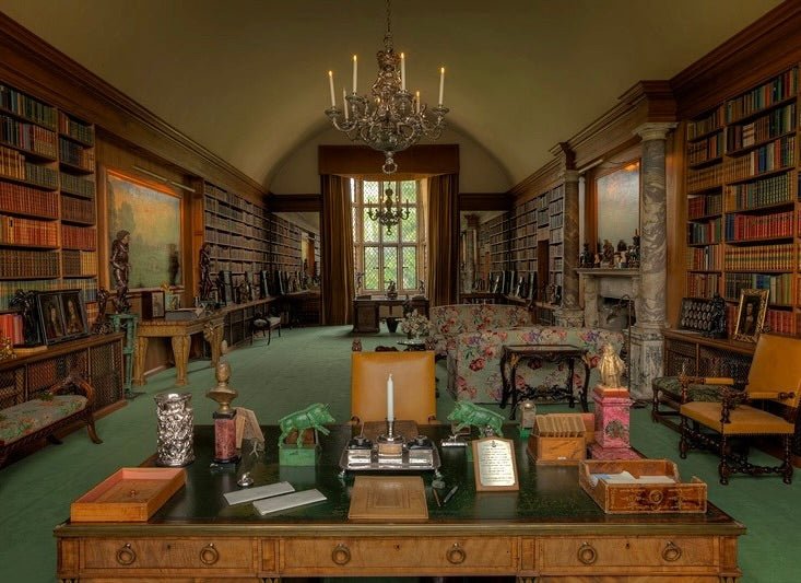 Elegant library with floor-to-ceiling bookshelves, ornate chandelier, wooden desk with candle, and green carpet throughout.