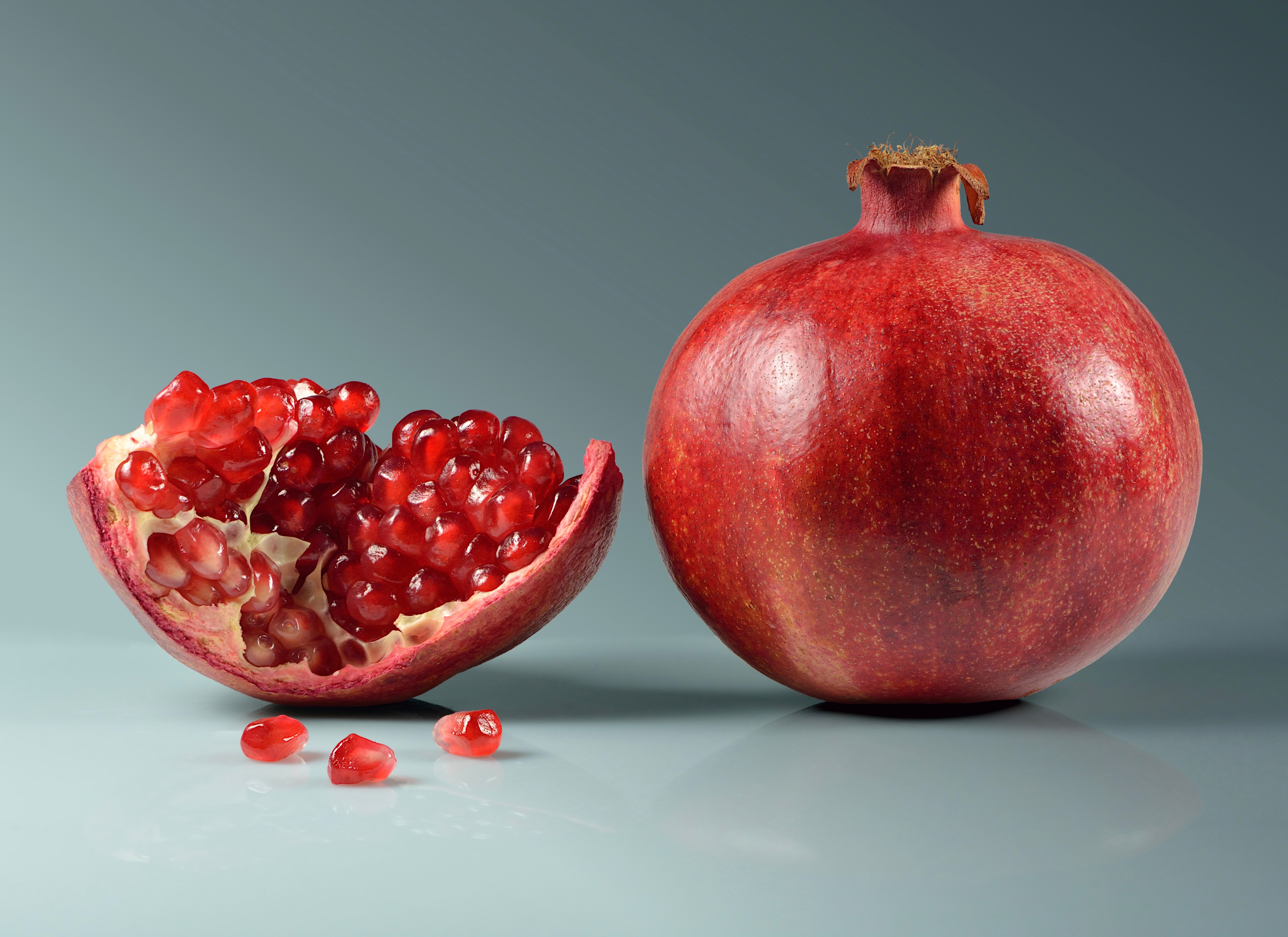 Whole red pomegranate next to halved section showing bright red arils inside, with scattered seeds on gray surface.