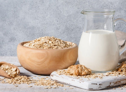 Wooden bowl of rolled oats with glass pitcher of white oat milk and oatmeal cookies on white wooden board.