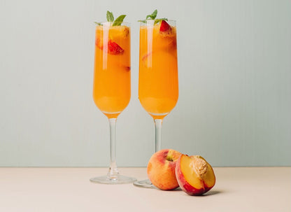 Two champagne flutes with orange peach Bellini cocktails garnished with mint and strawberry, with fresh peaches nearby.