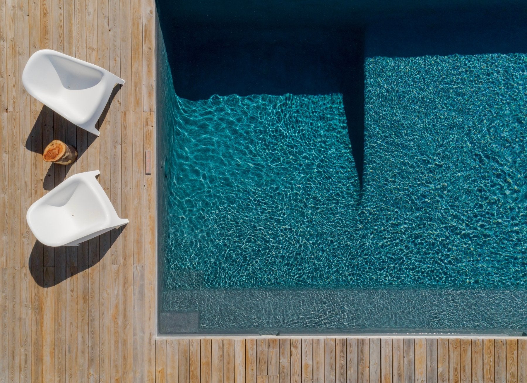 Aerial view of swimming pool with turquoise water and two white lounge chairs on wooden deck beside pool.