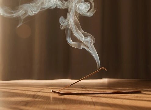 Burning incense stick on wooden surface with white smoke curling upward against blurred brown background.