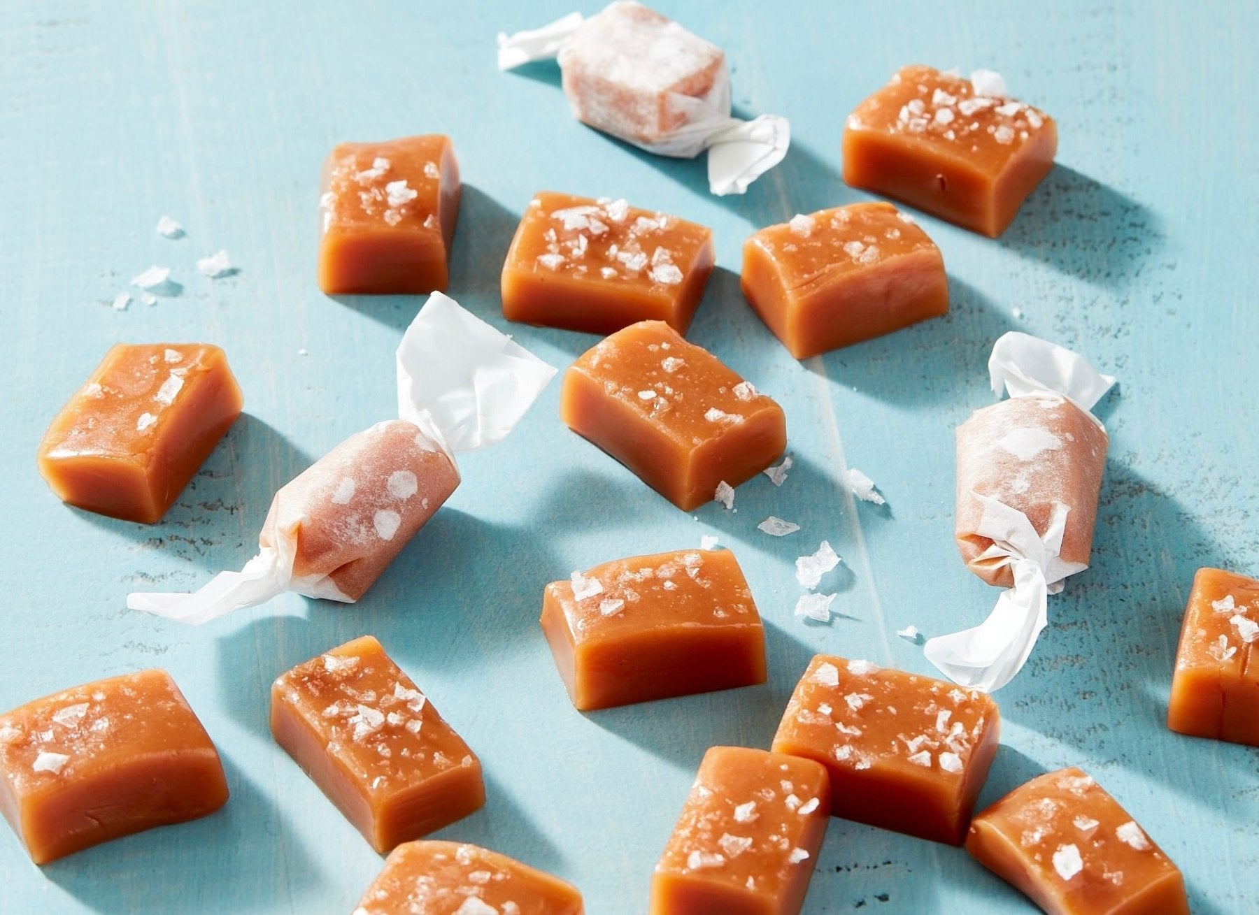 Golden caramel candies with white sea salt crystals scattered on light blue surface, some wrapped in clear cellophane.