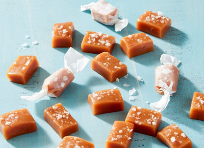 Golden caramel candies with white sea salt crystals scattered on light blue surface, some wrapped in clear cellophane.