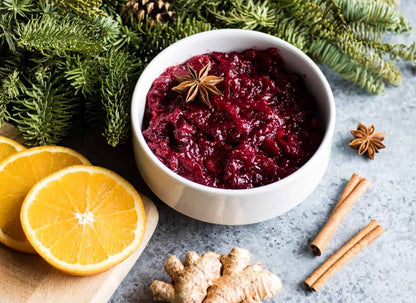 White bowl of red cranberry sauce with star anise garnish, surrounded by orange slices, cinnamon sticks, ginger, and evergreen branches.