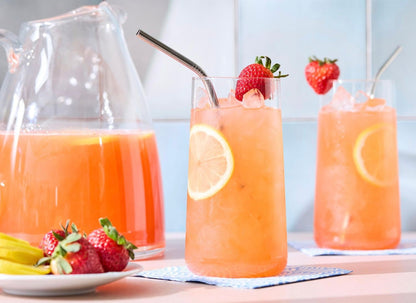 Orange-pink strawberry lemonade in tall glasses with lemon slices and strawberry garnish, with glass pitcher and fresh strawberries nearby.