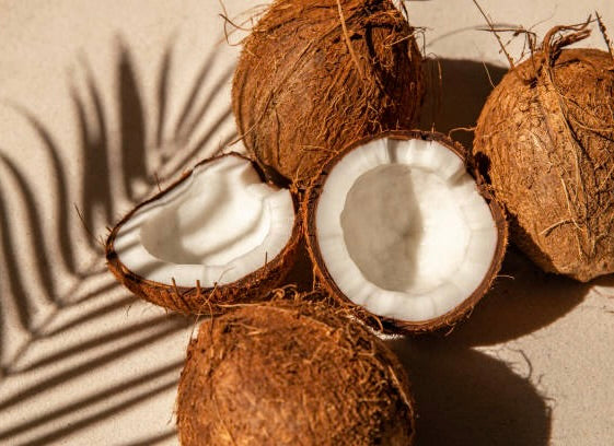 Brown coconuts with fibrous husks and one cracked open showing white flesh inside, with palm leaf shadows on beige surface.