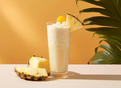 White creamy pina colada in tall glass with pineapple wedge garnish, fresh pineapple slices nearby, and tropical plant leaves in background.