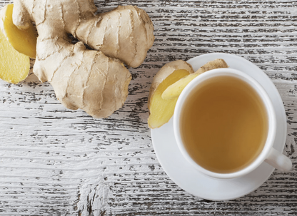White cup of golden tea with lemon slice garnish next to fresh ginger root with knobby texture on weathered white wood surface.